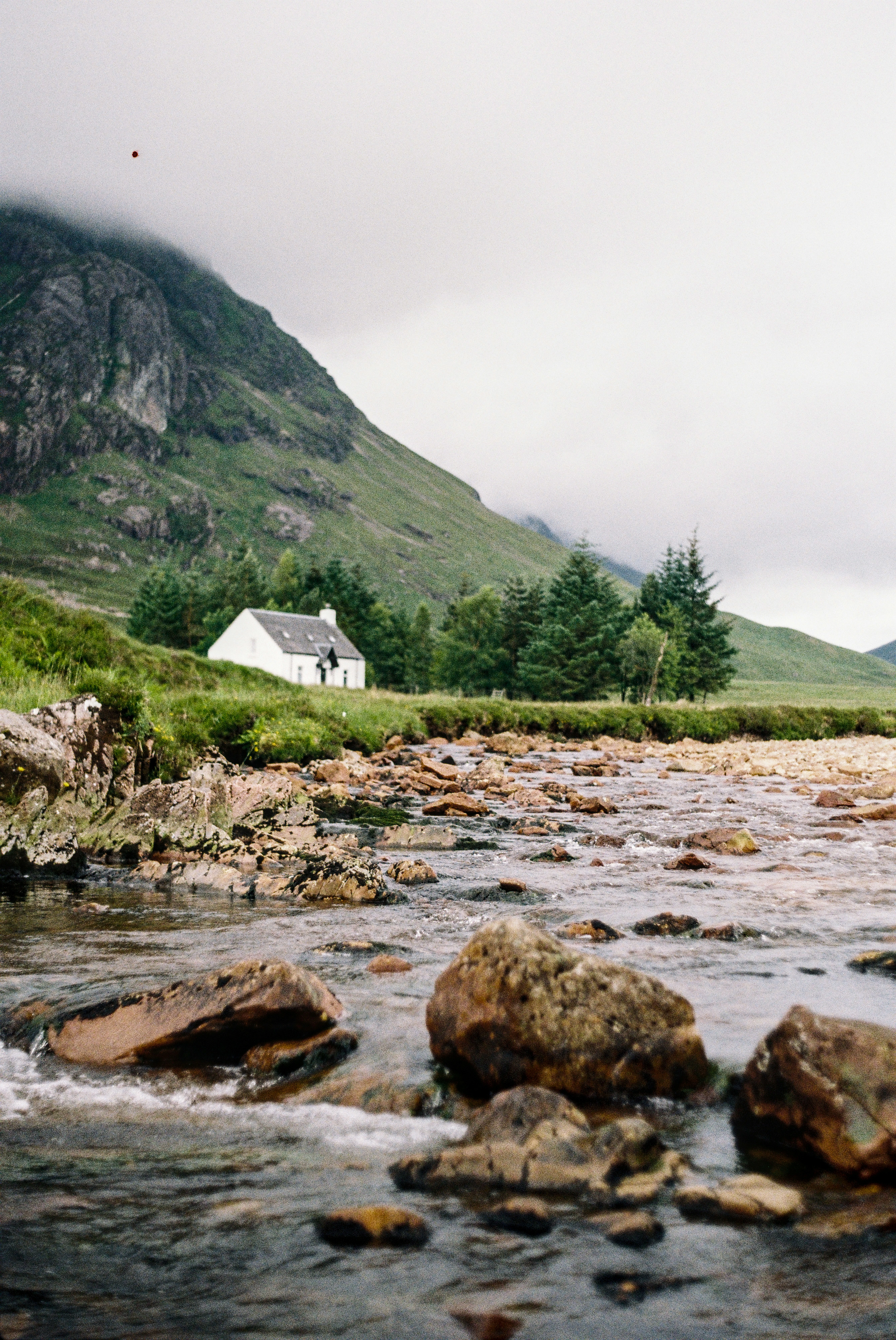 Et lille hvidt sommerhus beliggende ved siden af ​​en bæk omgivet af skovklædte bjerge
